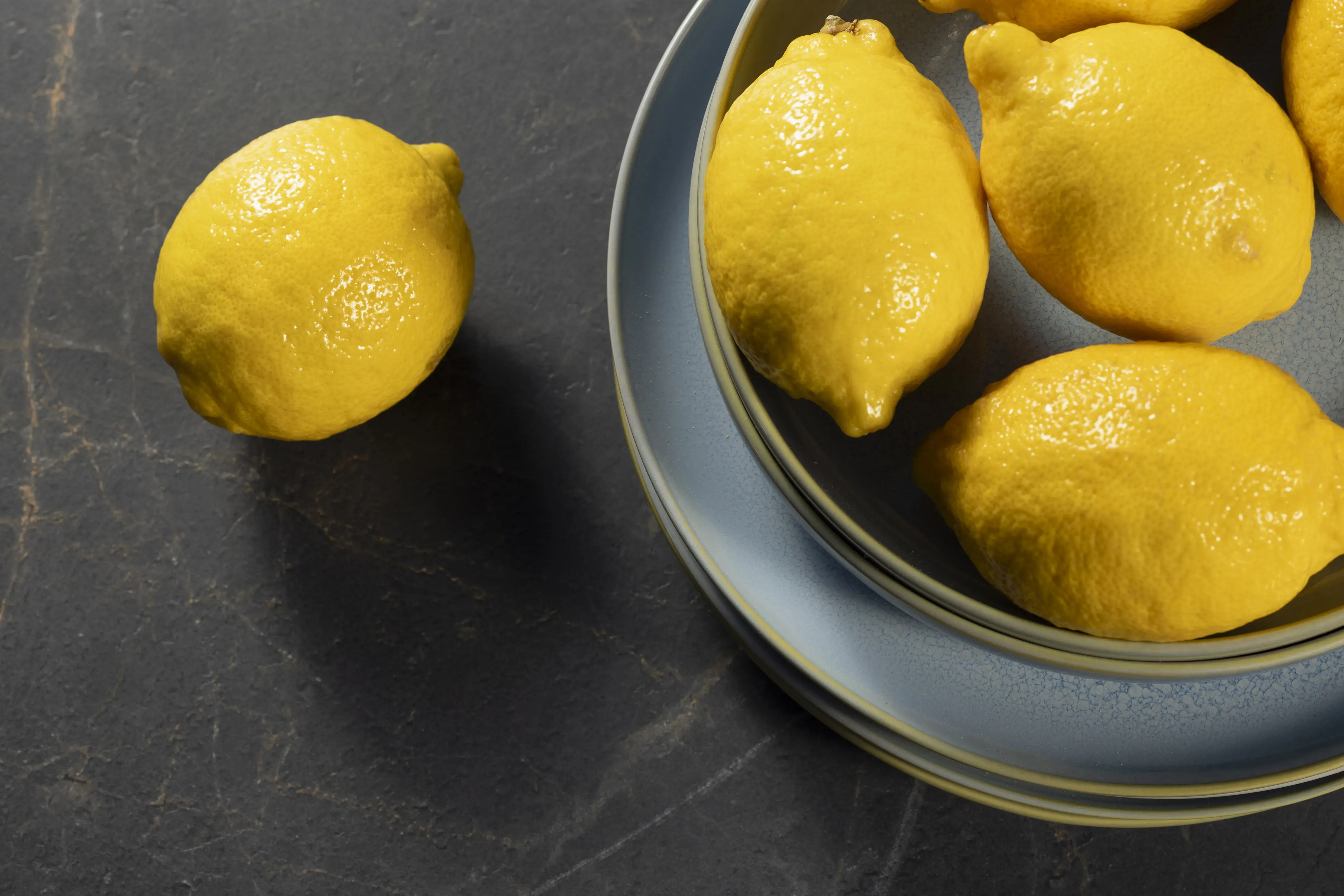 Fünf Zitronen auf einem blauen Teller, eine Zitrone daneben auf dunklem Untergrund.